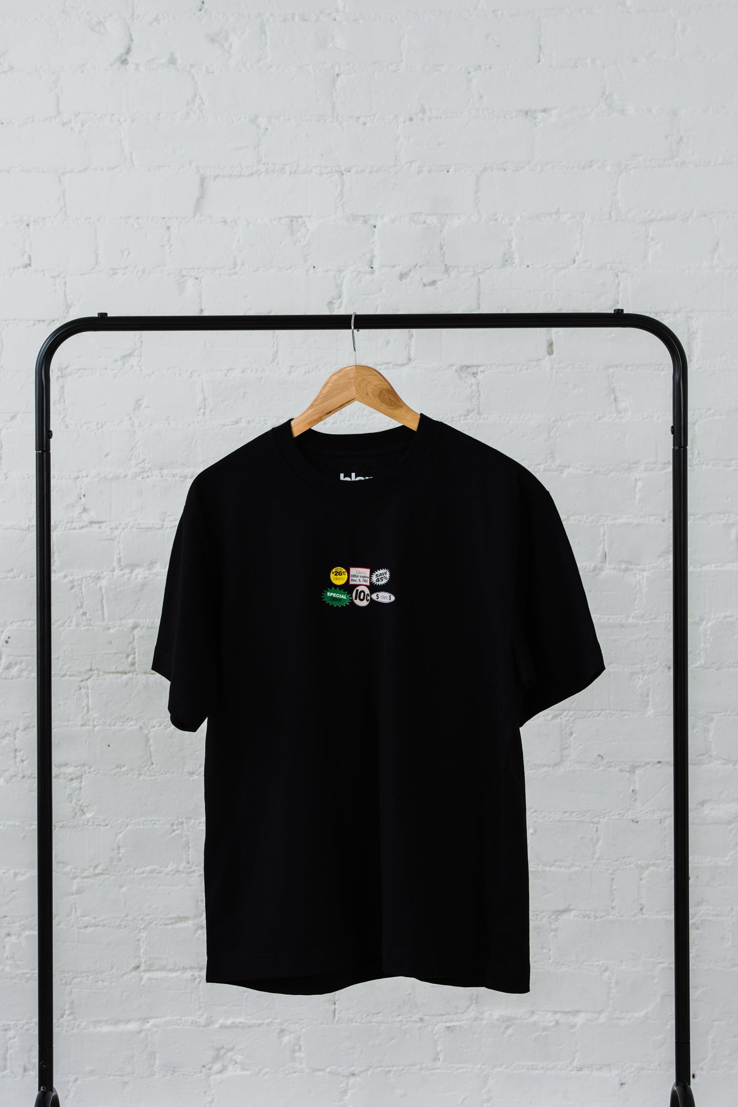 Black blanc t-shirt with colorful graphic on a hanger against a white brick wall.